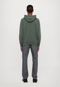 Felpa verde con cappuccio, dal taglio rilassato, con polsini e orlo a coste, abbinata a jeans grigi a gamba dritta e sneakers nere.