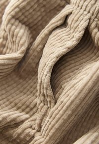 Beżowy materiał corduroy z subtelnymi pionowymi prążkami, miękką teksturą i złożonymi sekcjami, eksponującymi zaokrąglone krawędzie i organiczne kształty.