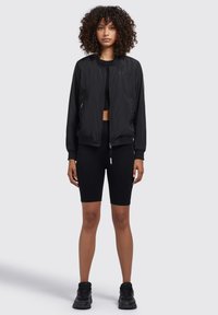 Jachetă bomber neagră cu manșete și talie ribbată, asortată cu un top scurt negru și pantaloni scurți de bicicletă. Material texturat și design minimalist.