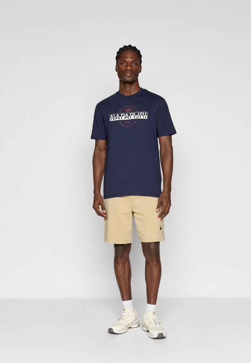 T-shirt bleu marine à manches courtes avec un design de logo blanc, associé à un short beige et des sneakers claires. Tenue simple et décontractée.