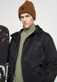 Giacca nera impermeabile con tasca frontale, sopra un pile verde con colletto alto, abbinata a un berretto di maglia marrone a trama. Snowboard sullo sfondo.