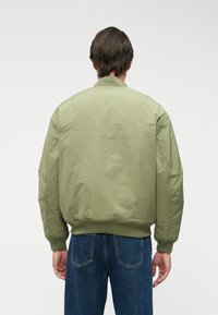 Πράσινο μπουφάν bomber με λεία υφή, ribbing στους καρπούς και το στρίφωμα, και χαμηλωμένα ραμφάκια στους ώμους. Φοριέται με μπλε τζιν παντελόνι.