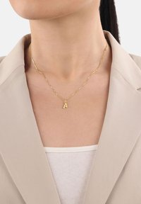 Persona che indossa una collana d'oro con un ciondolo a forma di "A", un blazer beige e un top bianco a coste, accorciato al collo e al petto.