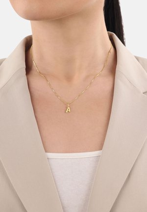 Person, die eine goldene Halskette mit einem "A"-Anhänger, ein beigefarbenes Blazer und ein weißes, geripptes Oberteil trägt, das am Hals und an der Brust gekürzt ist.