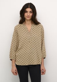Beige blouse met lange mouwen en zwarte stippen, V-halslijn en drie knopen aan de voorkant. Gathered mouwen zorgen voor een ontspannen pasvorm.