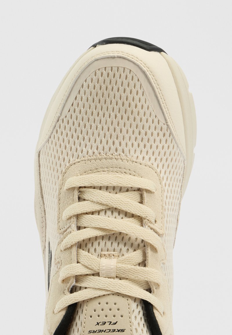 Zapatilla deportiva beige con empeine de malla transpirable, cordones texturizados, cuello acolchado y un acento negro en la puntera. Logotipo de Skechers en la suela.