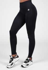 Black leggings with a high waistband, featuring a textured pattern and a small logo, paired with white and black athletic shoes.