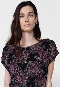 Haut noir à manches courtes avec broderie florale dans des tons de rose et de gris, accentué par un col en perles. Texture de tissu douce.