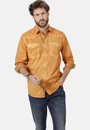 Oranges, strukturiertes Hemd mit Knöpfen und zwei Brusttaschen, hochgekrempelten Ärmeln und kontrastierenden Innensaumdetails. Kombiniert mit dunklen Jeans.