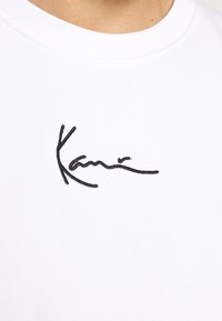 Camiseta de algodón blanca con un diseño de firma bordado en negro en el frente. Cuello redondo, mangas cortas, textura suave.