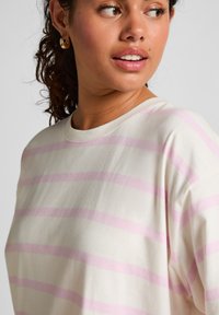 Vrouw met krullend haar, die gouden oorringen en een wit shirt met lichtroze horizontale strepen draagt, kijkend opzij.