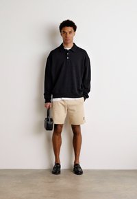 Svart pullover med tre knappar, beige shorts och svarta loafers. Håller en liten svart handväska. Enkelt, avslappnat outfit.