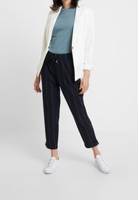 Blazer blanco sobre una camisa verde azulado, combinado con pantalones rayados azul marino y zapatillas blancas. El blazer tiene botones dorados y los pantalones tienen una cinturilla elástica.
