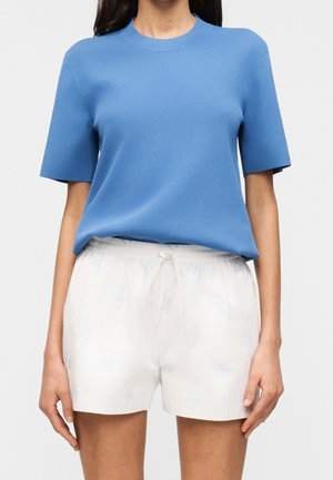 Femme portant un haut en maille bleu clair à manches courtes et un short blanc avec cordon de serrage, avec un motif subtil sur un fond uni.