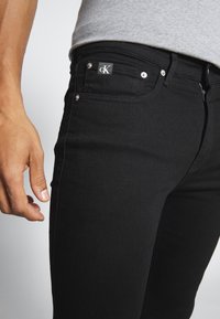 Primer plano de una persona que lleva puesta unos pantalones vaqueros negros de Calvin Klein con el logo visible en el bolsillo y una camiseta gris claro metida por dentro.