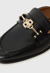 Mocassino in pelle nera con finitura liscia, dotato di un accento in hardware color oro e dettagli cuciti sulla parte superiore.