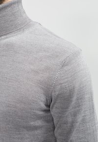 Pull à col roulé gris en tricot avec une texture lisse, une coupe ajustée et des détails subtilement côtelés au niveau du col et des coutures.