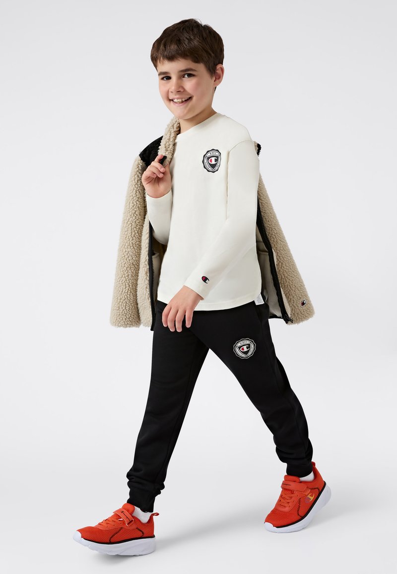 Biała koszula z długim rękawem z czarnym logo, czarne dresy z logo oraz czerwone sneakersy. Na sobie ma beżową kurtkę z owczej wełny.