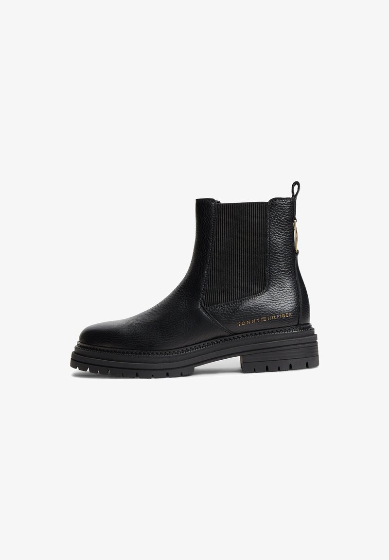 Bottine noire en cuir avec panneau latéral élastique, languette à l’arrière, semelle épaisse à crampons et petit logo doré Tommy Hilfiger sur le côté.