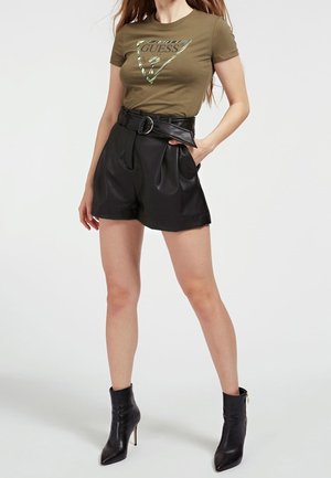Woman wearing olive green Guess t-shirt, high-waisted black leather shorts with belt, and black high-heeled ankle boots, standing.