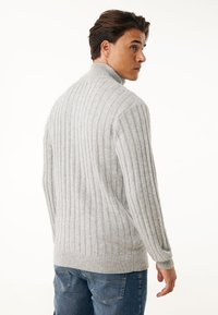 Helles graues geripptes Sweatshirt mit hohem Kragen und langen Ärmeln, aus weichem, strukturiertem Stoff. Getragen mit blauen Denim-Jeans.