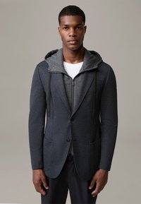 Ciemnoszara marynarka z teksturowanej tkaniny, z szarą bluzą z kapturem na zamek błyskawiczny pod spodem, kontrastowymi wzorami kieszeni i akcentami w postaci sznurka.