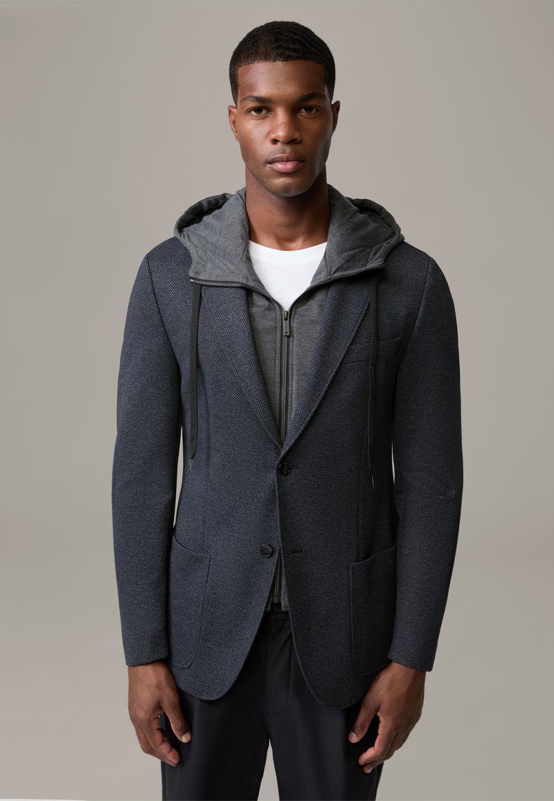 Ciemnoszara marynarka z teksturowanej tkaniny, z szarą bluzą z kapturem na zamek błyskawiczny pod spodem, kontrastowymi wzorami kieszeni i akcentami w postaci sznurka.