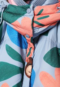 Farverig jakke med en lyseblå bund, prydet med store grønne og koralfarvede blomsterprint, blød tekstur og et bjørneansigt nær knapperne.