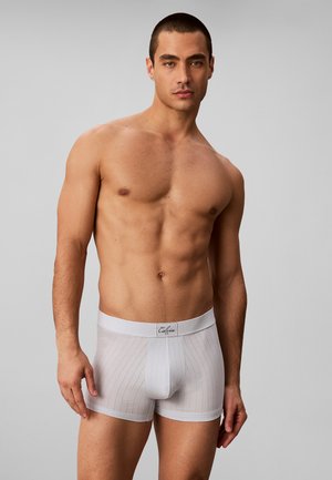 Muskuløs mand med kort hår iført hvide stribede boxershorts med et mærketaljebånd, stående mod en ensfarvet grå baggrund.
