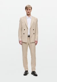 Abito beige chiaro a doppio petto con texture fine, vestibilità su misura e pantaloni abbinati. Camicia bianca sotto; cintura e scarpe nere.