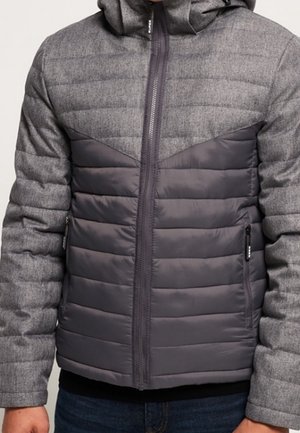 Homme portant une veste à capuche grise avec des sections matelassées et rembourrées, une fermeture éclair à l'avant et des poches latérales zippées.