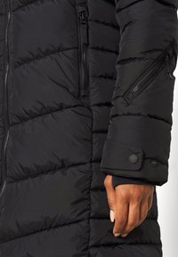 Abrigo tipo plumífero negro con diseño acanalado, que cuenta con un bolsillo lateral con cremallera y detalle de puño abotonado en la manga. Tejido suave y texturizado.