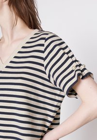 Top de cuello en V rayado con franjas horizontales beige y azul marino. Manga corta con bordes con volantes y botones de acento en perla en los puños. Textura de tela suave.