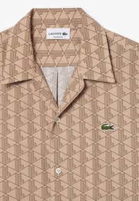 Brązowa, wzorzysta koszula z krótkim rękawem o geometrycznym wzorze. Posiada zapięcie na guziki oraz haftowane zielone logo krokodyla na klatce piersiowej.