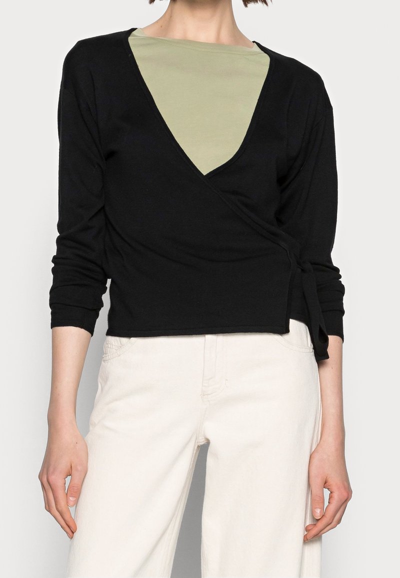Personne portant un pull cache-cœur noir sur un haut vert clair et un pantalon blanc, debout devant un arrière-plan neutre.