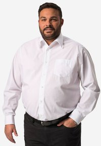 Camicia bianca a maniche lunghe con colletto classico, taschino anteriore singolo e maniche con polsini, abbinata a pantaloni scuri e una cintura nera.