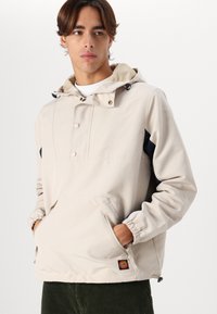 Beige hooded jacket with navy side panels, featuring a structured design, snap buttons, elastic cuffs, and a front pocket. Fanta Cruz logo visible.