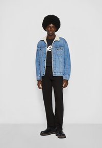 Hombre con afro que lleva una chaqueta de mezclilla azul con forro de borrego, camiseta gráfica negra, pantalones negros y botas negras, de pie frente a un fondo gris.