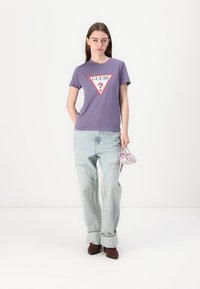 Lilla t-skjorte med en rød og hvit trekantet "GUESS"-logo. Kombinert med lyseblå baggy-jeans og brune ankelstøvler, holder en liten lilla veske.