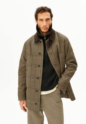 Mann mit kurzen lockigen Haaren, der eine braune karierte Jacke über einem schwarzen Pullover und beigen Hosen trägt, steht vor einem weißen Hintergrund.