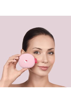 Frau mit klarer Haut hält eine pinke Hello Kitty Gesichtsbürste nahe an ihrer Wange vor einem hellvioletten Hintergrund.