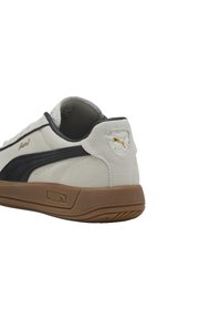 Baskets blanches et noires avec une tige en daim, semelle en caoutchouc marron, accents de logo dorés et un patch sur le talon texturé comportant un logo.