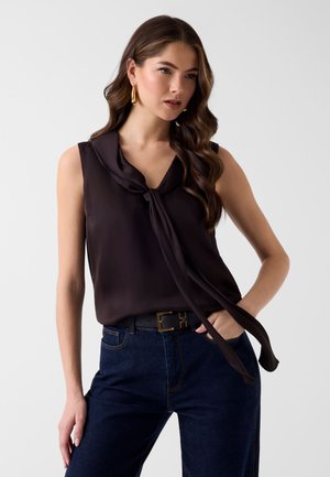 Femme aux longs cheveux ondulés portant un haut noir sans manches avec un col à nouer, des boucles d'oreilles créoles dorées, un jean bleu foncé et une ceinture noire avec une boucle dorée.