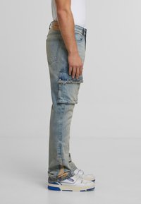 Homme portant un jean cargo en denim bleu clair déchiré avec un rabat de poche effiloché et des baskets blanches avec des accents bleus, se tenant de côté.