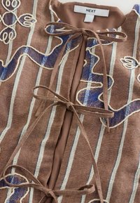 Vêtement en lin marron avec des rayures verticales blanches et bleues, motifs floraux brodés, et trois nœuds attachés au niveau de l'encolure devant.