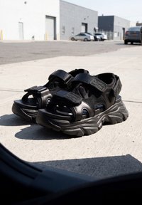 Ζευγάρι μαύρα chunky πλατφόρμα σανδάλια με λουριά βέλκρο που τοποθετούνται σε ηλιόλουστο τσιμεντένιο πεζοδρόμιο κοντά σε σταθμευμένα αυτοκίνητα και κτίρια.