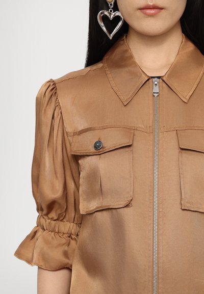 Braune Satinbluse mit Reißverschluss vorne, kurzen Puffärmeln, zwei Brusttaschen und plissierten Akzenten. Große silberne herzförmige Ohrringe.