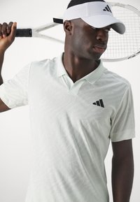 Polo shirt texturé vert clair avec un logo noir, porté avec une visière blanche. Une raquette de tennis est tenue sur l'épaule.