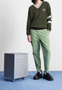 En person klädd i grön Nike-tröja och byxor med svarta klumpiga skor står bredvid en metallisk rullande förvaringslåda på ett blått golv.