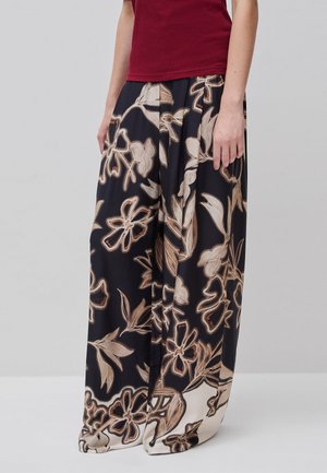 Person trägt lockere schwarze Hosen mit weitem Bein und beigen Blumenmustern sowie ein bordeauxrotes Oberteil, steht vor einem hellgrauen Hintergrund.